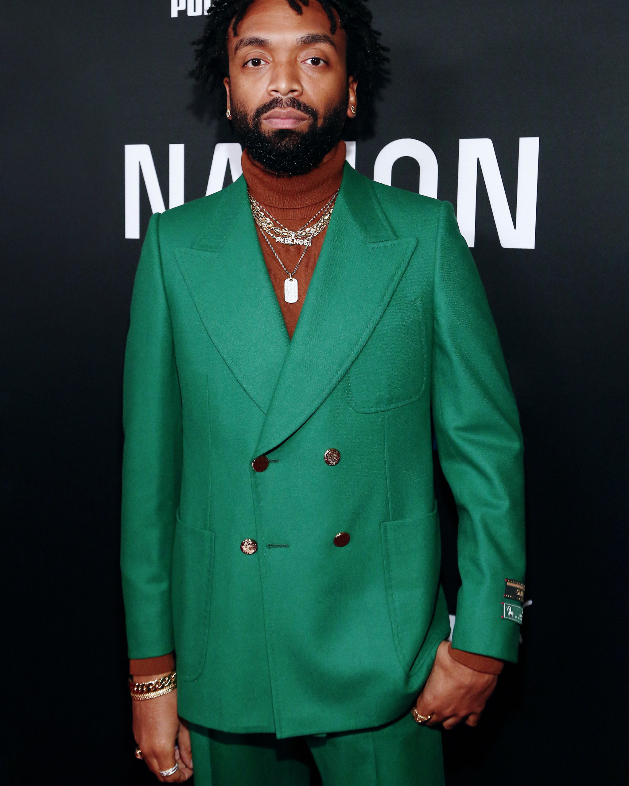 Kerby Jean-Raymond at the 2020 Roc Nation Brunch wearing a kelly green suit and a selection of retro jewelry including a set of gold chain necklaces and custom diamond stud earrings from Shelley & Co
